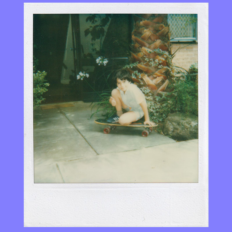 Photo of Sean as a very small child sitting, crouched on a vintage skateboard next to plants and a tree.
    He is wearing shorts and blue sneakers.
